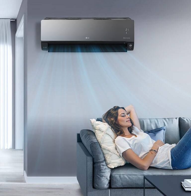 A woman lounges on a couch in a living room with the LG air conditioner installed above her on the wall. Blue streams of air are on the image to indicate it is on and cooling the room.