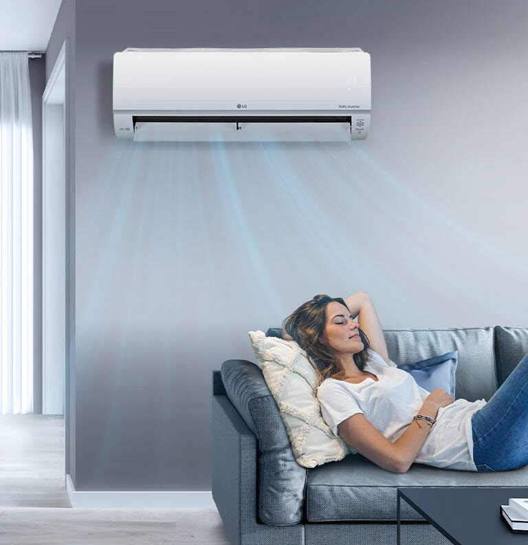 A woman lounges on a couch in a living room with the LG air conditioner installed above her on the wall. Blue streams of air are on the image to indicate it is on and cooling the room.