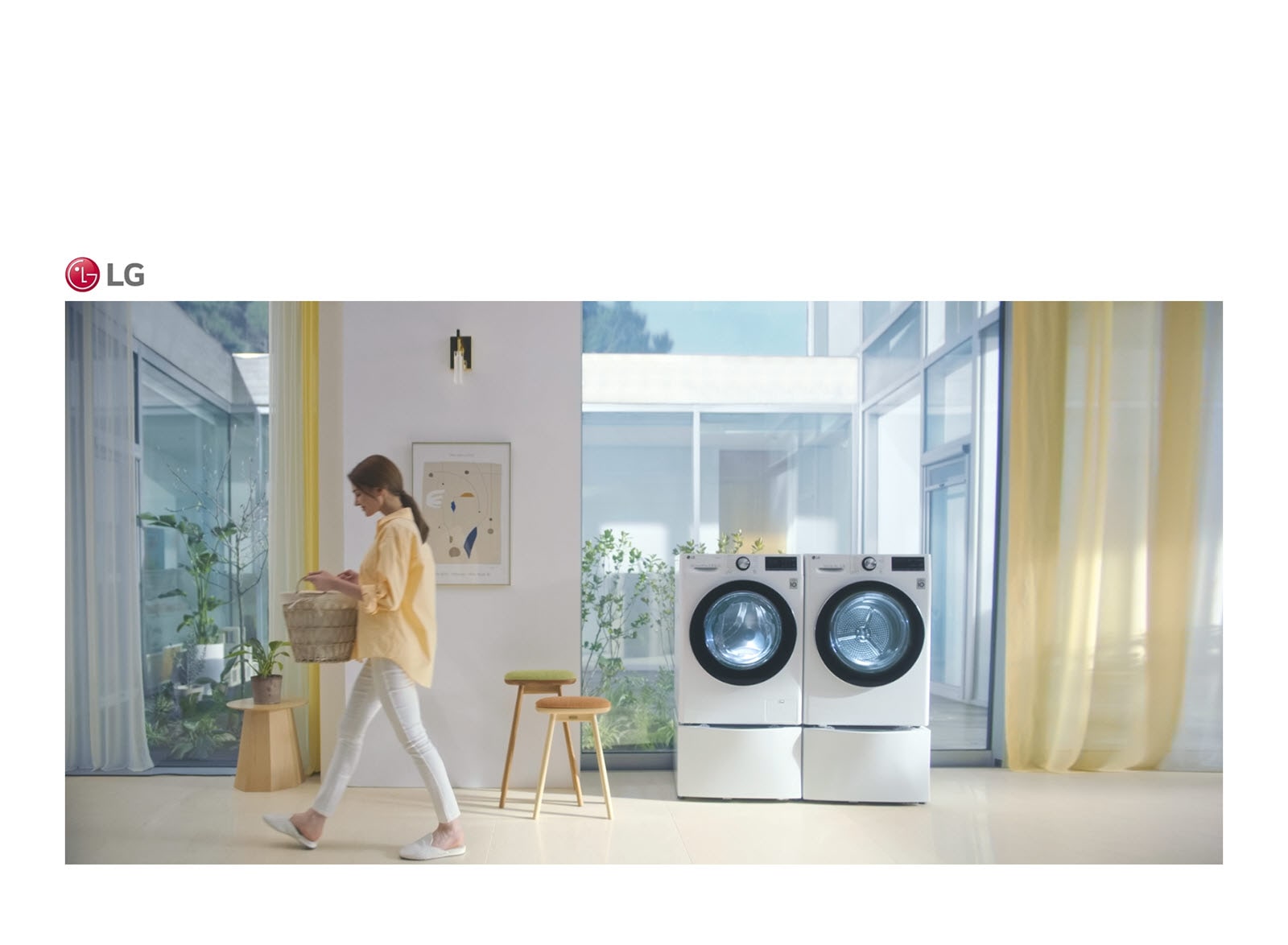 A washing machine is at the center of the room. A woman walks by with a basket of laundry.