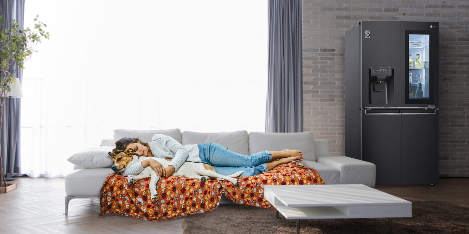 A refrigerator sits against a brick wall with a sofa to the side and a woman and dog sleeping peacefully on the sofa.