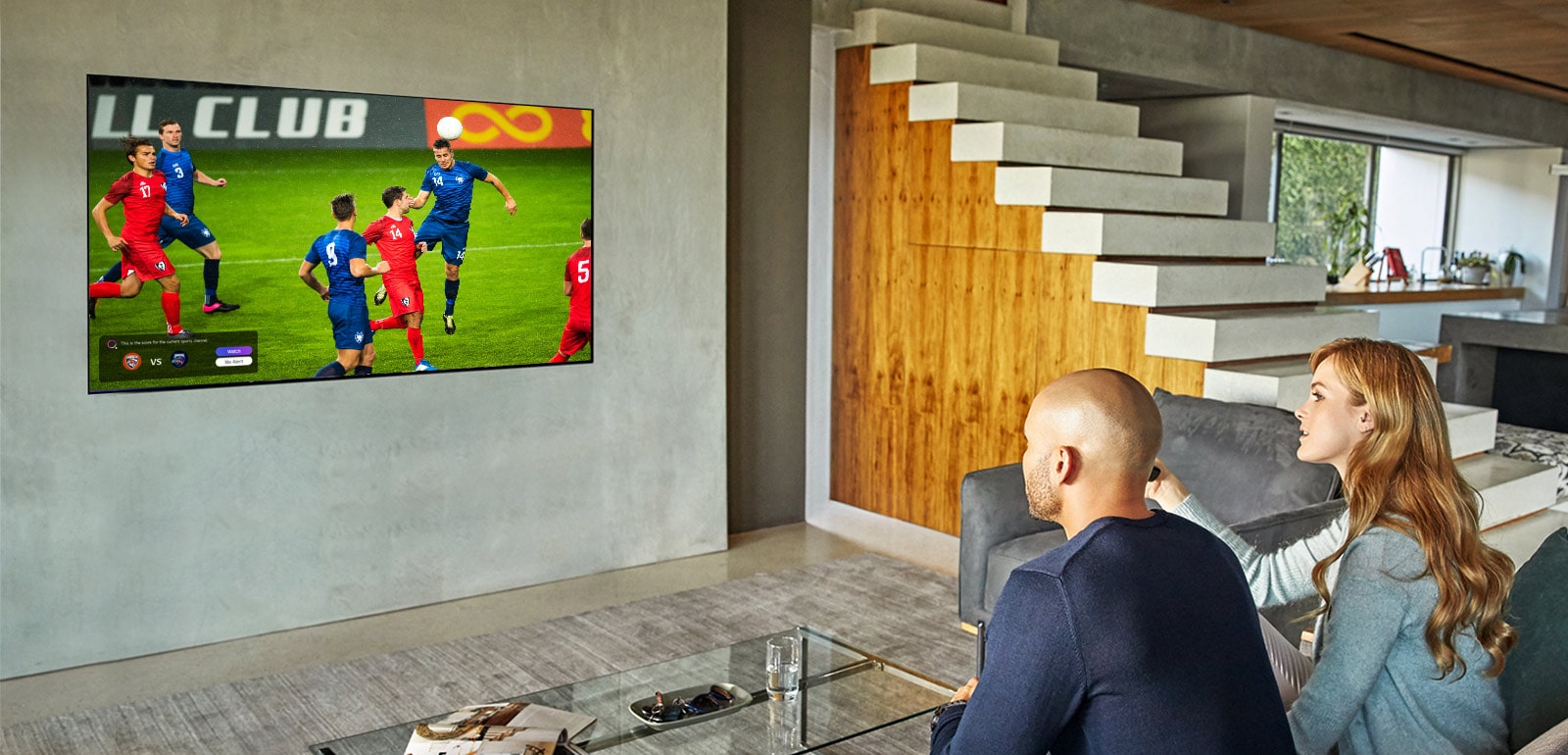 A man and woman sat behind a coffee table in front of a wall mounted TV showing a football match.