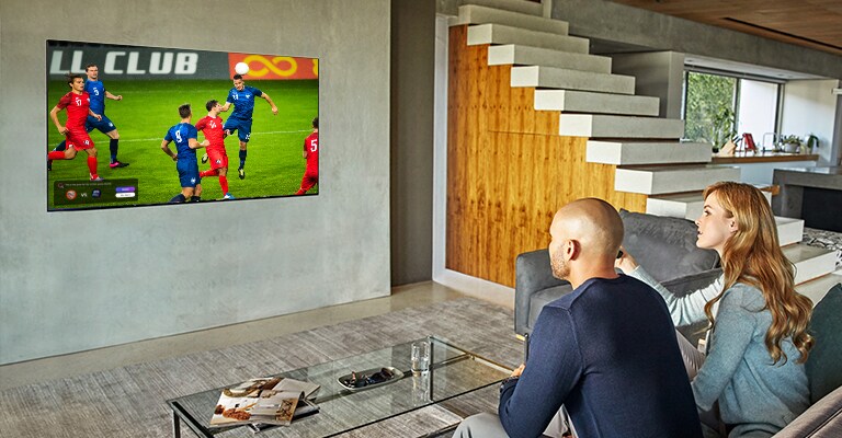 A man and woman sat behind a coffee table in front of a wall mounted TV showing a football match.