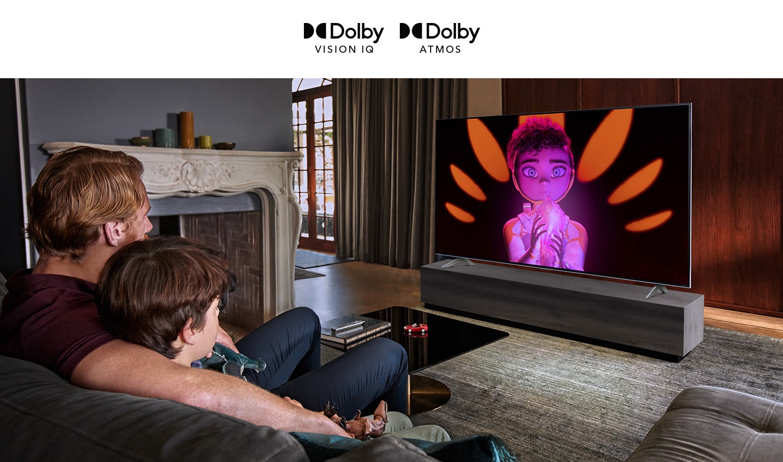 The logo of Dolby Vision IQ and Atmos are in line horizontally. Under the logos, a father and a son sitting on a couch are watching television displaying  a girl  holding a mineral in the middle of black and orange background.