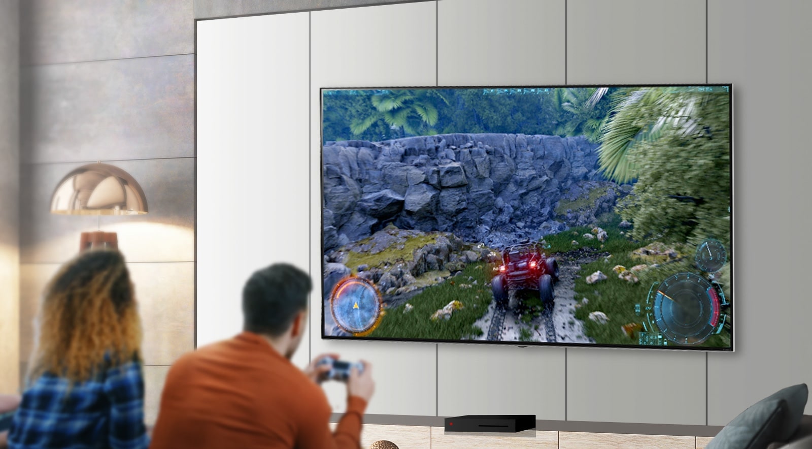 Rear view of a man and woman sat side-by-side in front of a large wall-mounted TV. The man is holding a gaming controller with a driving game on screen.