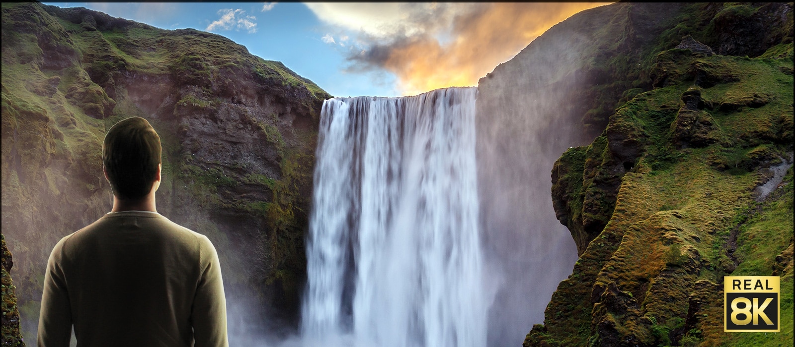 A man stood in front of an imposing view of a large waterfall crashing down cliffs. The scene zooms out to show the waterfall as an image on a wall mounted TV.