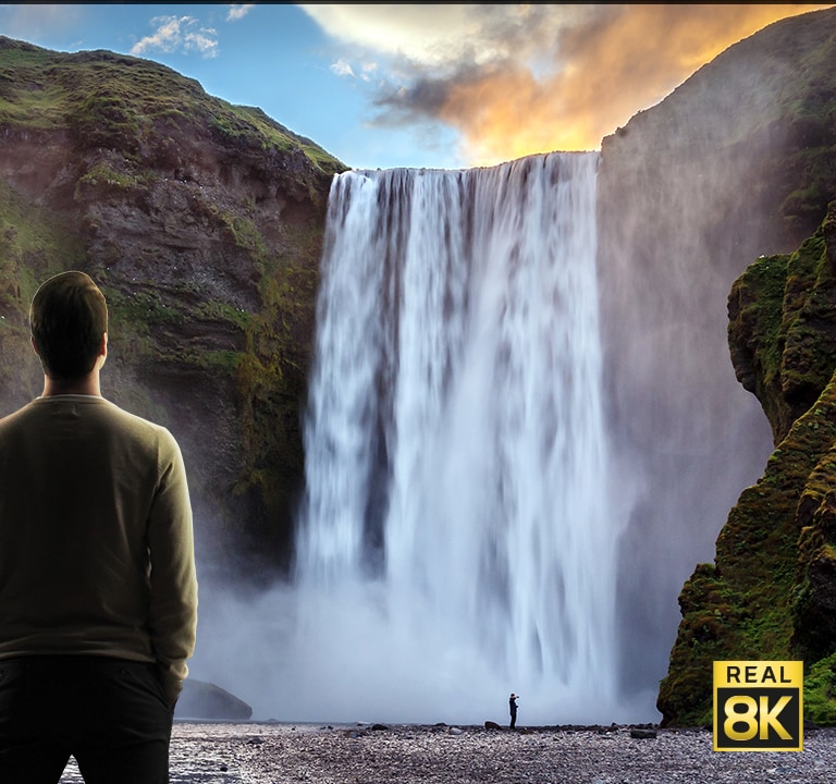 A man stood in front of an imposing view of a large waterfall crashing down cliffs. The scene zooms out to show the waterfall as an image on a wall mounted TV.