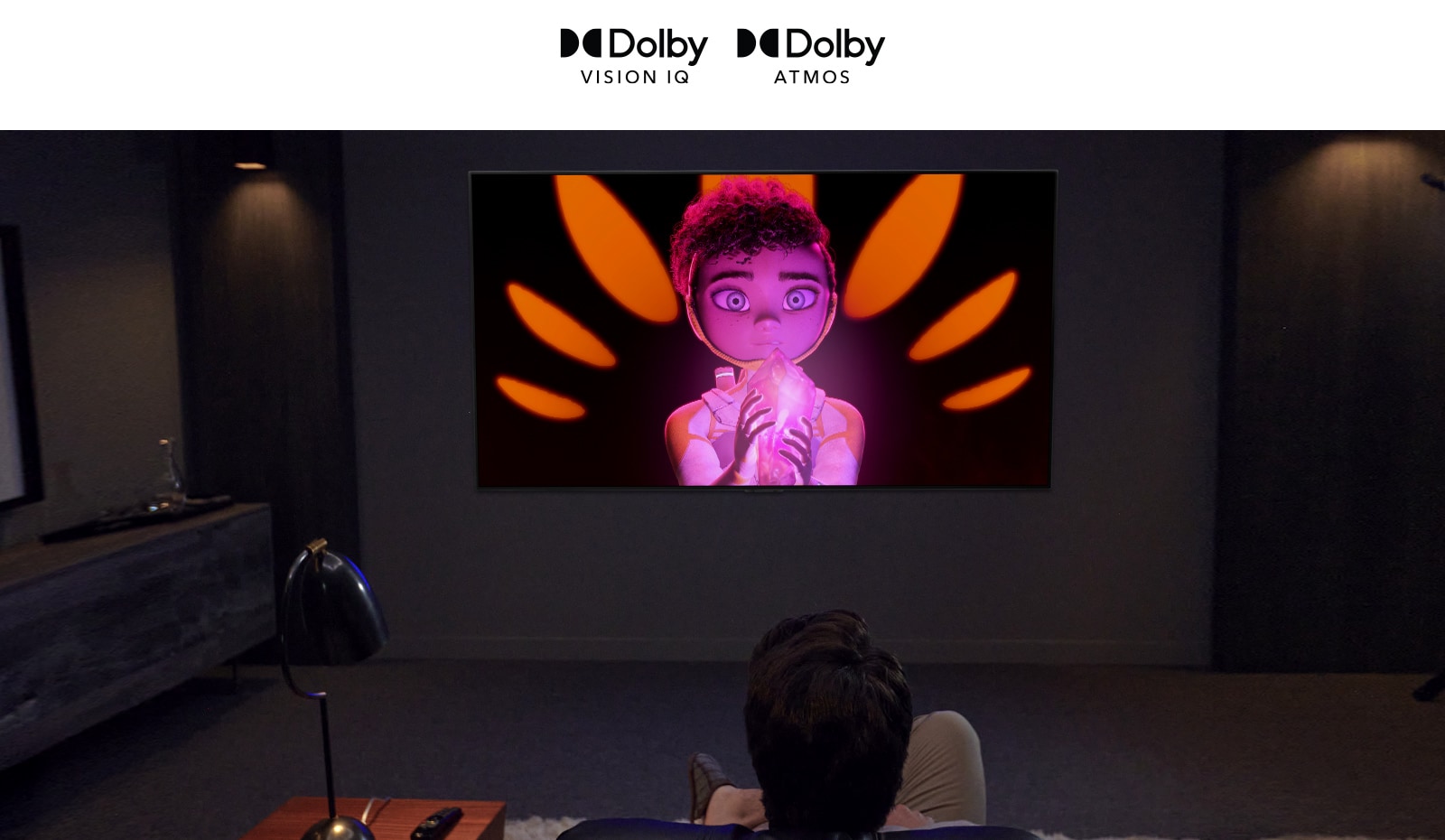 The logo of Dolby Vision IQ and Atmos are in line horizontally. Under the logos, a father and a son sitting on a couch are watching television displaying  a girl  holding a mineral in the middle of black and orange background.